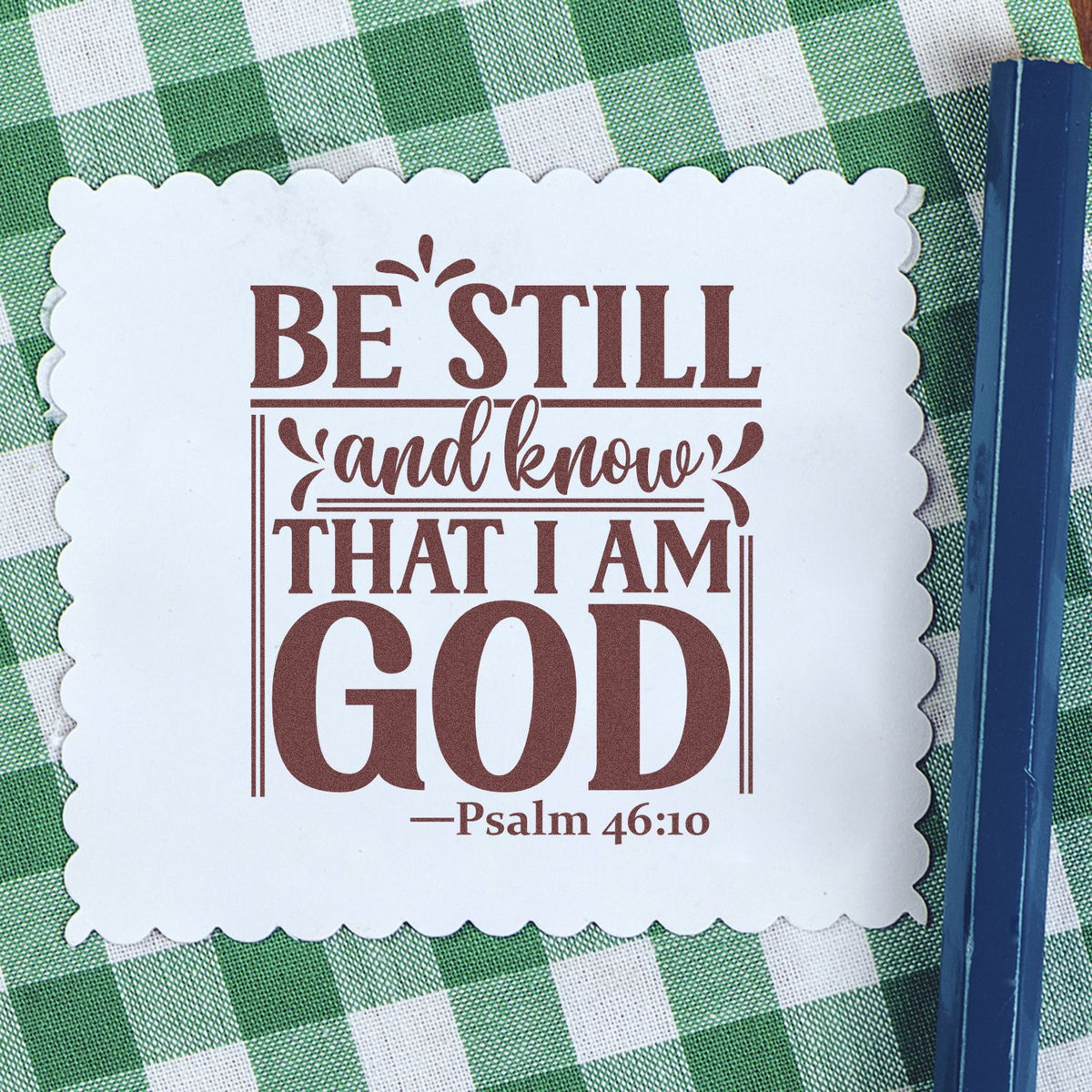 Scalloped white stamp seal with brown Psalm 46:10 text on green gingham, beside a blue stamping tool.