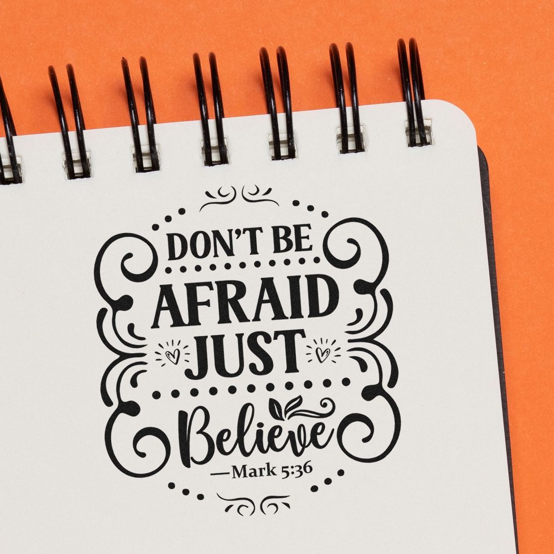 Black decorative stamp and seal design on white notebook page with orange background, reading Dont Be Afraid Just Believe Mark 5:36.