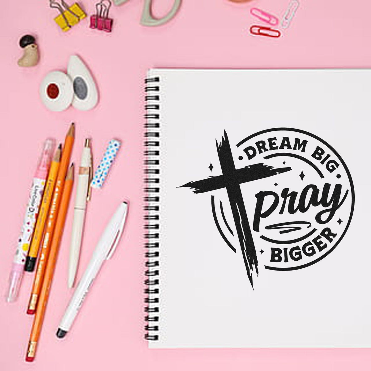 Black dream-big-pray-bigger stamp seal on a white spiral notebook, with pink stationery and pens on a pastel pink background.