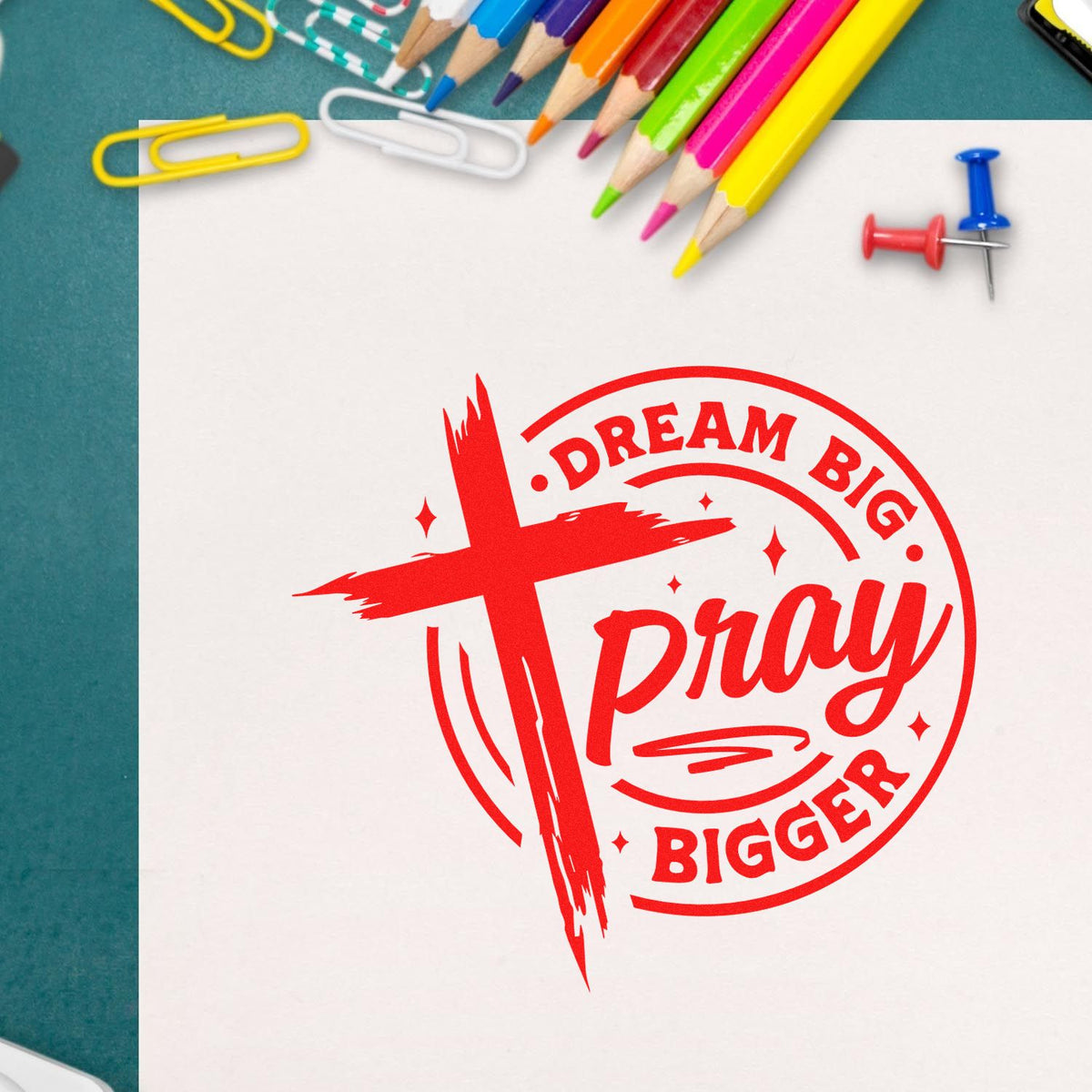 Red Dream Big Pray Bigger stamp and seal design on white paper, with colorful pencils, paperclips, and teal background.