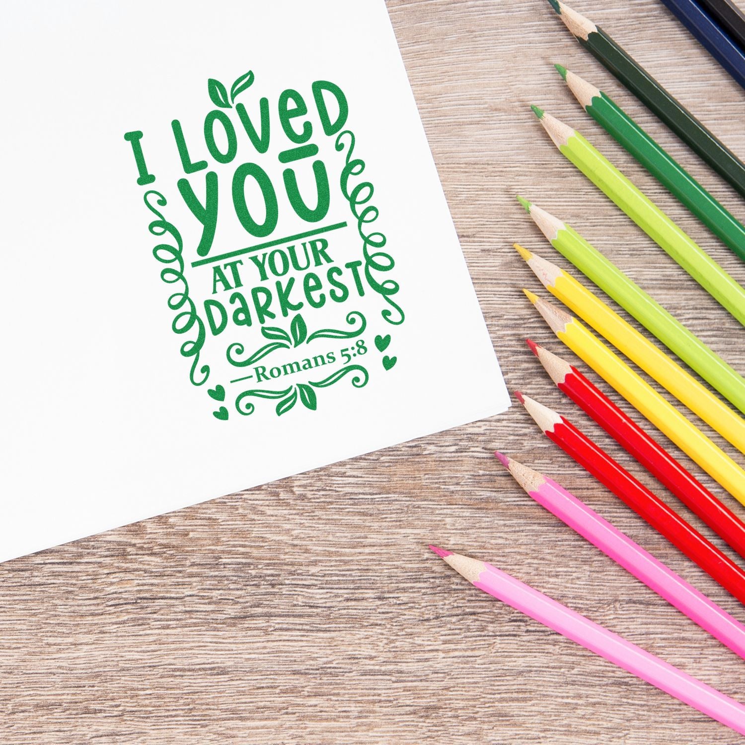 Green decorative stamp seal with Bible verse I Loved You at Your Darkest beside colorful pencils on a wood desk.