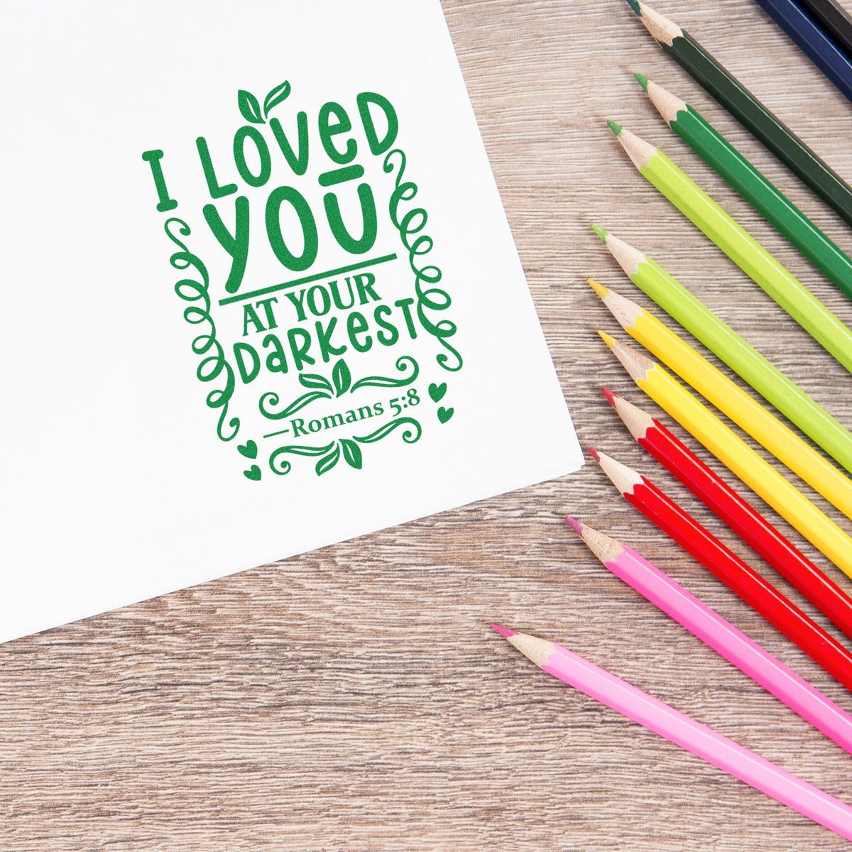 Green stamp and seal reading I Loved You at Your Darkest with Romans 5:8, on white paper beside colorful pencils.
