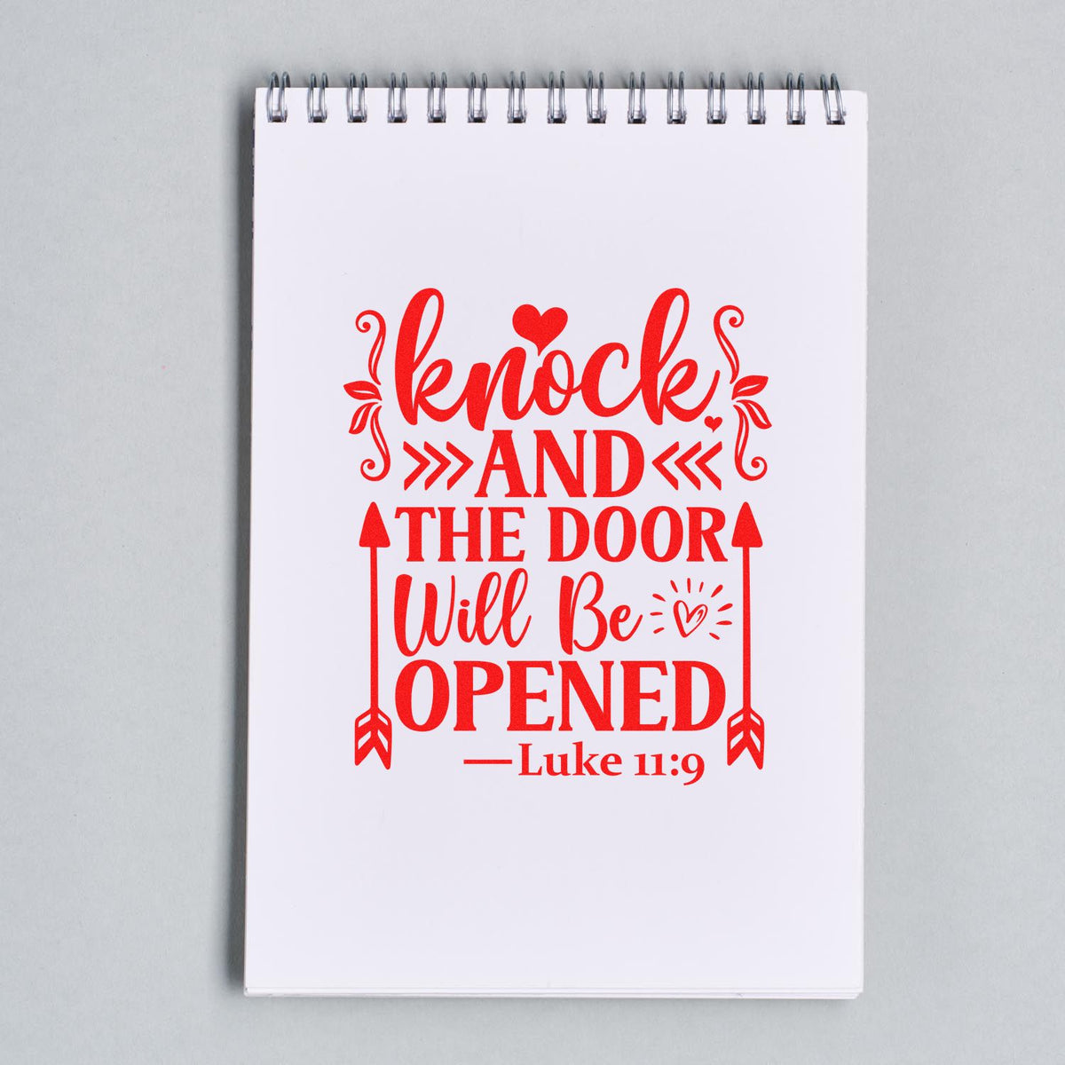 Red decorative stamp and seal design on a white spiral notebook reading Knock and the door will be opened Luke 11:9.