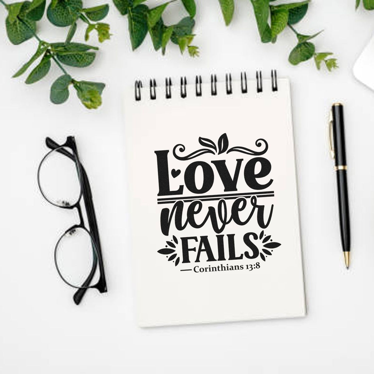 Black Love never fails Corinthians 13:8 stamp and seal design on a white notebook, with green leaves, glasses, and pen.