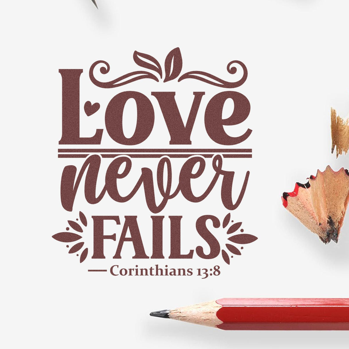 Maroon decorative stamp/seal reading Love Never Fails with Corinthians 13:8, floral accents, on white with pencil tip and shavings.