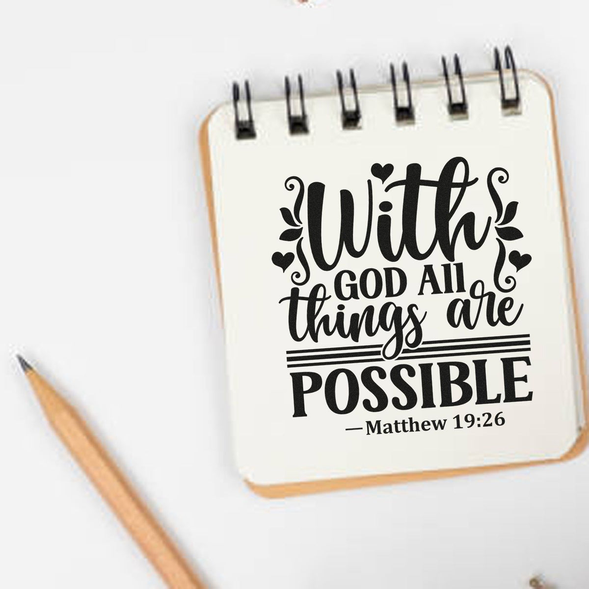 White spiral notebook stamp with black scripture seal design reading With God all things are possible, beside a pencil.