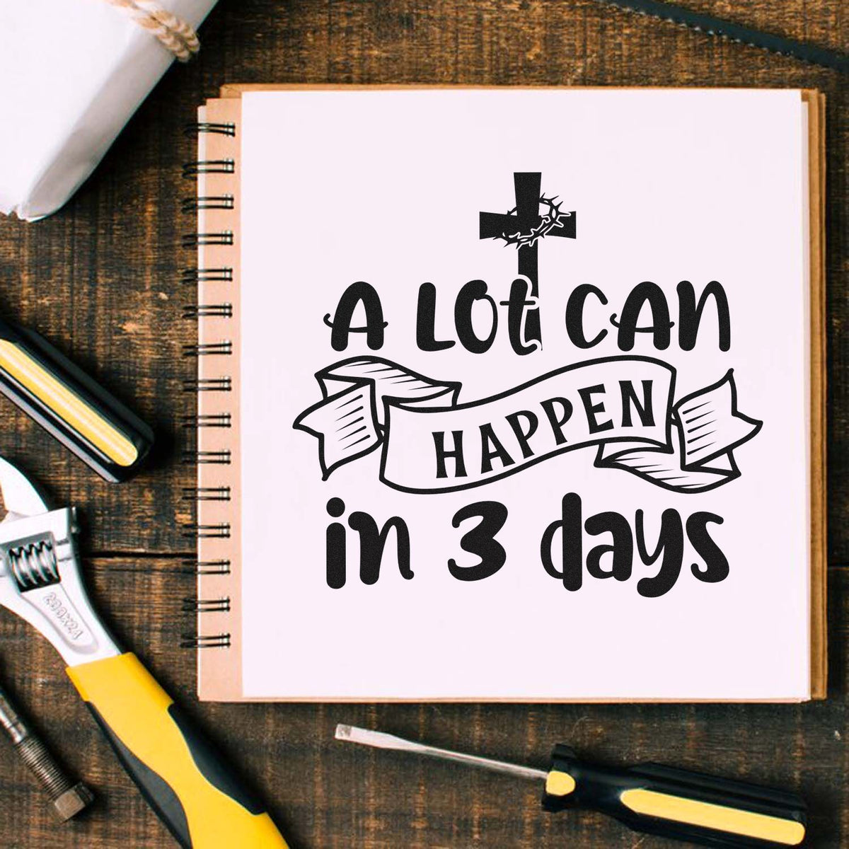 Black church stamp and seal design with cross and A lot can happen in 3 days on a white notebook, beside yellow-and-black tools.