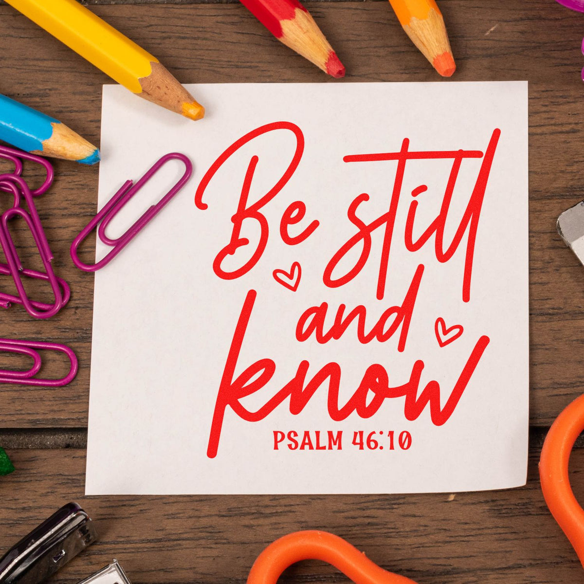 Red stamp/seal design reading Be still and know with Psalm 46:10 on white paper, framed by colorful pencils and purple paper clips.