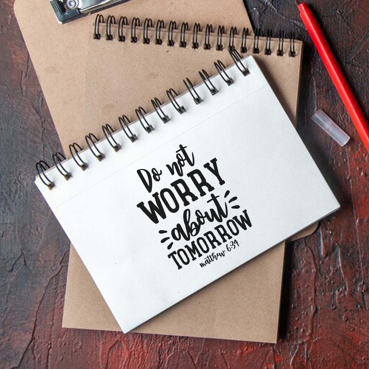 Custom black stamp on white spiral notebook, stacked over a kraft notebook with a red pen on dark wood.