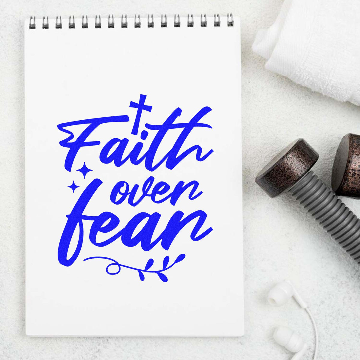 Blue Faith over fear script on a white notebook beside stamp and seal tools, with white fabric and earbuds.