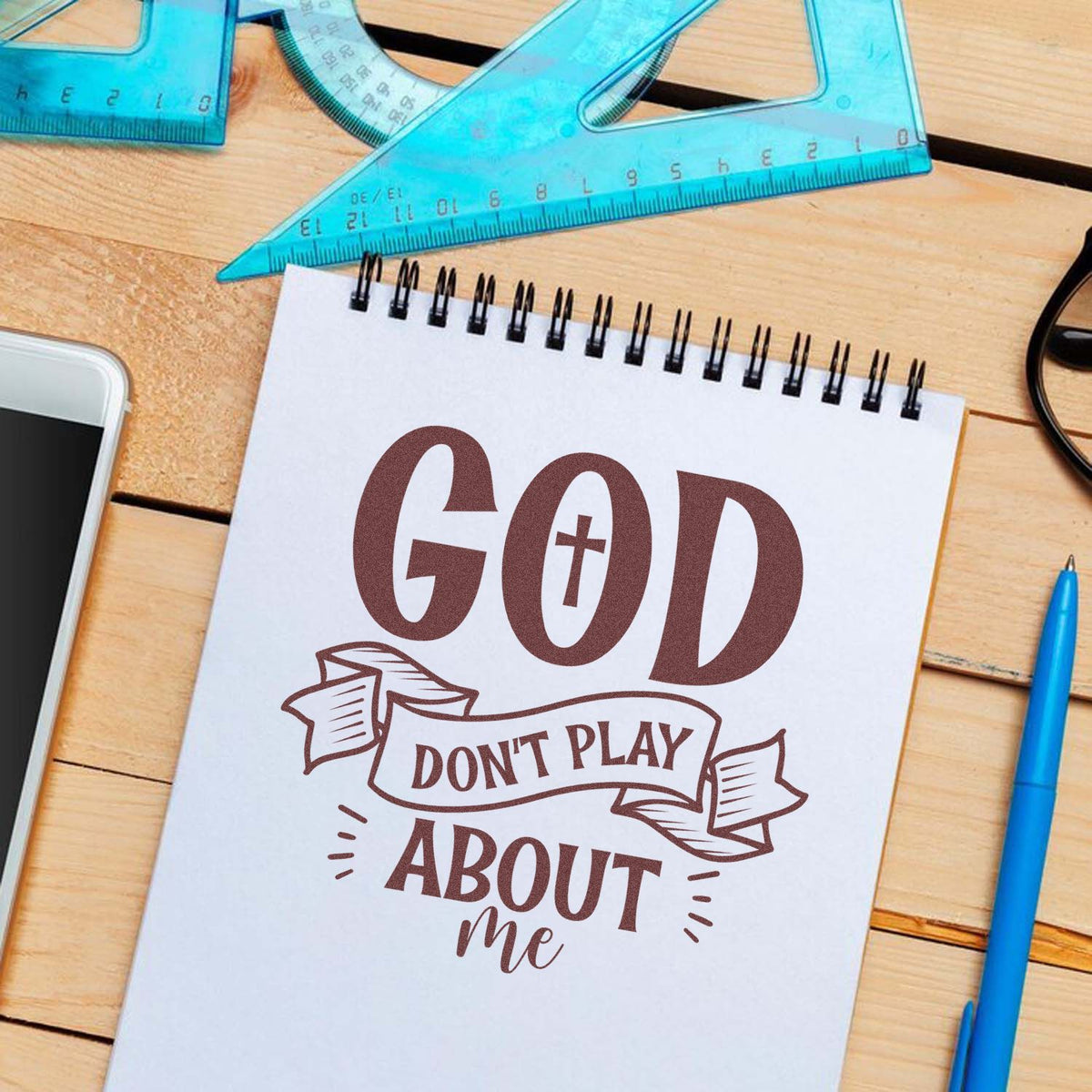 Brown faith-themed rubber stamp and seal reading God dont play about me on white notebook, with blue rulers and pen.
