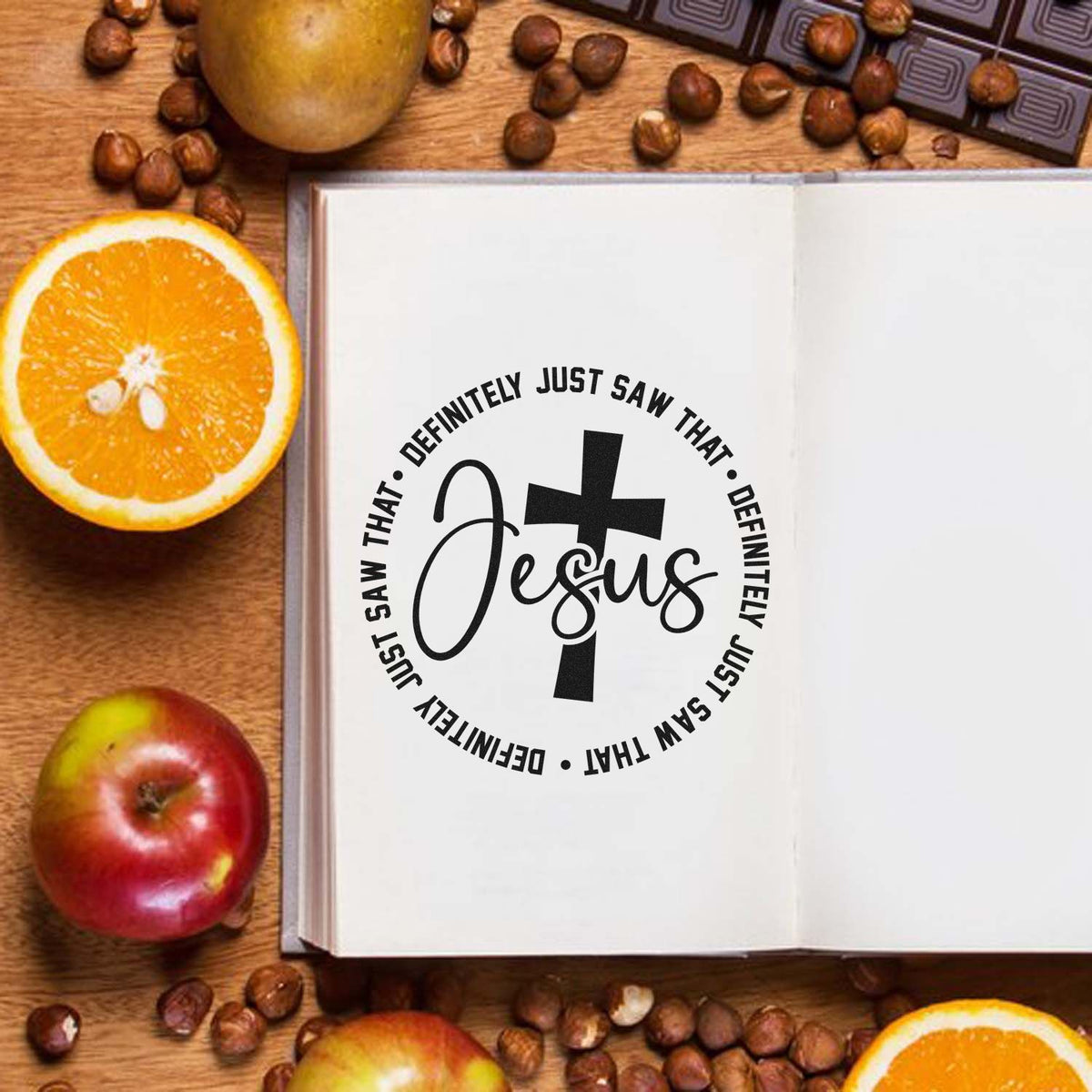Black Jesus cross stamp seal on open white book, framed by oranges, apples, and coffee beans on warm wood.