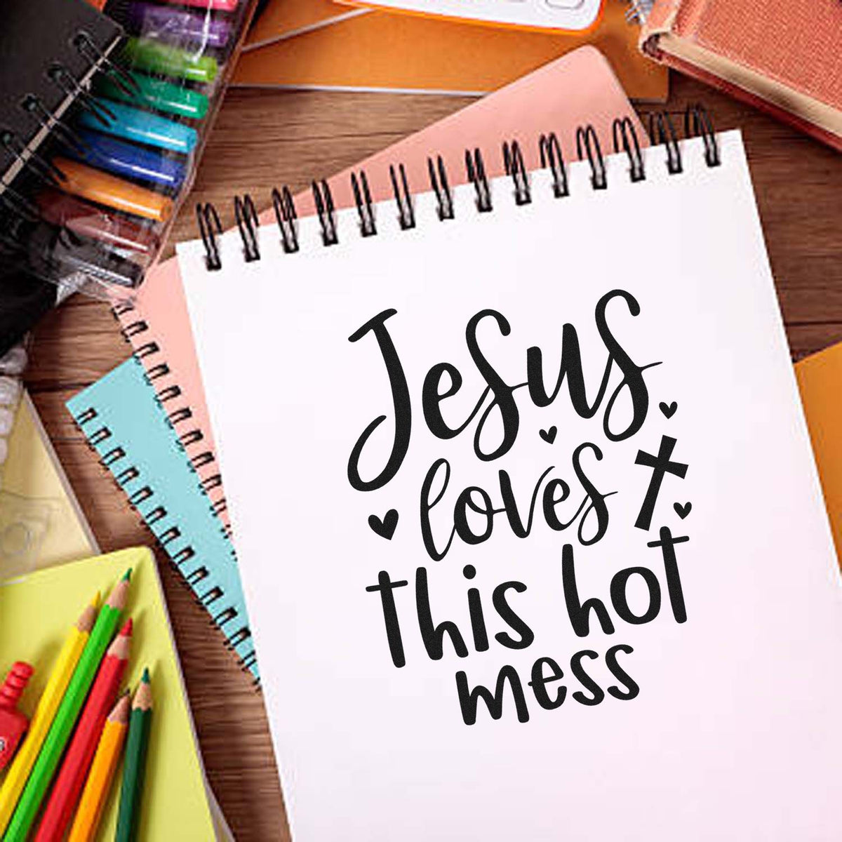 White spiral notebook with black Jesus loves this hot mess stamp-style seal, surrounded by colorful pens and pastel notebooks.
