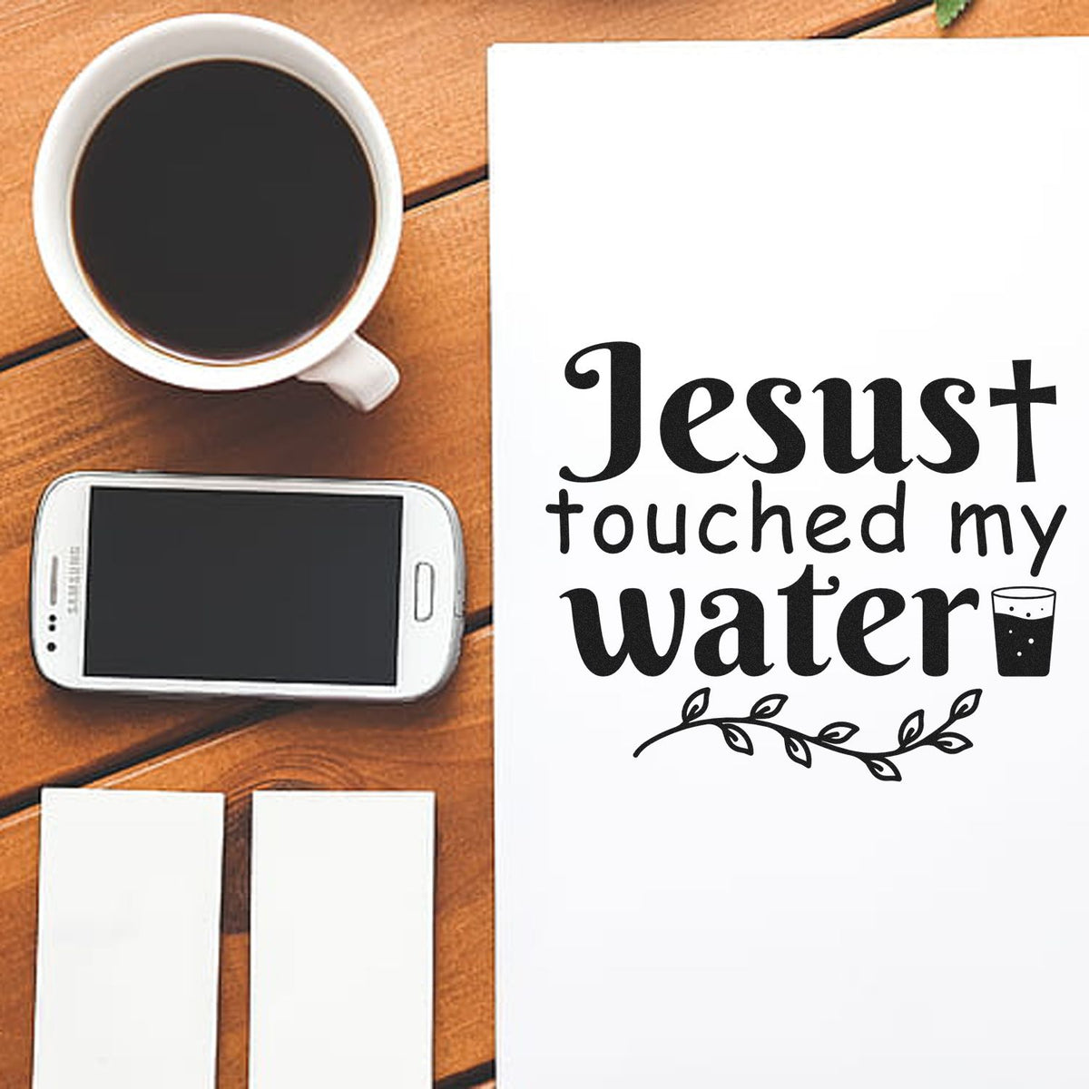 White stamp and seal products with black Jesus touched my water design, on a wood table beside coffee and a phone.