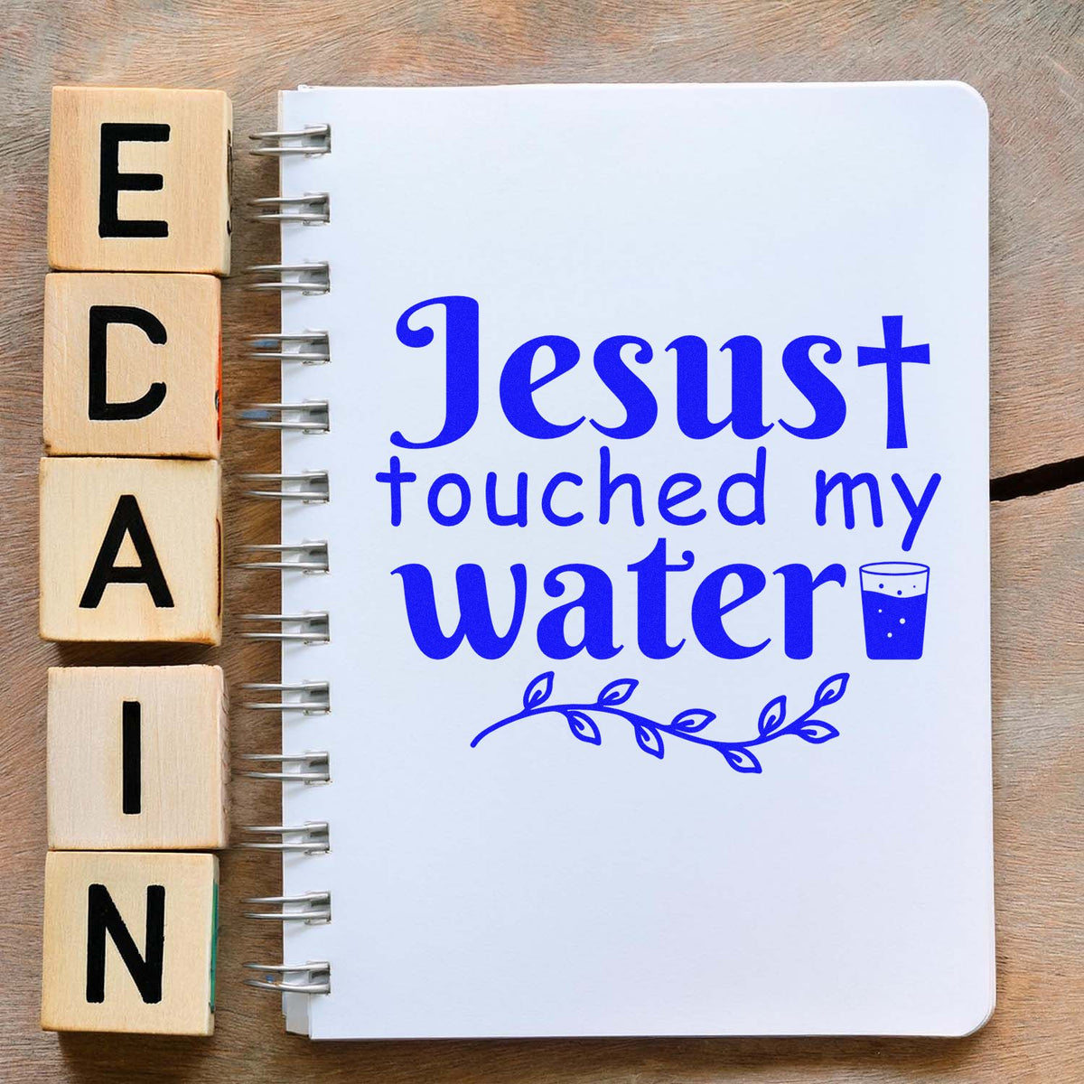 Wooden stamp blocks beside a spiral notebook with blue Jesus touched my water text and cup icon on a wood surface.