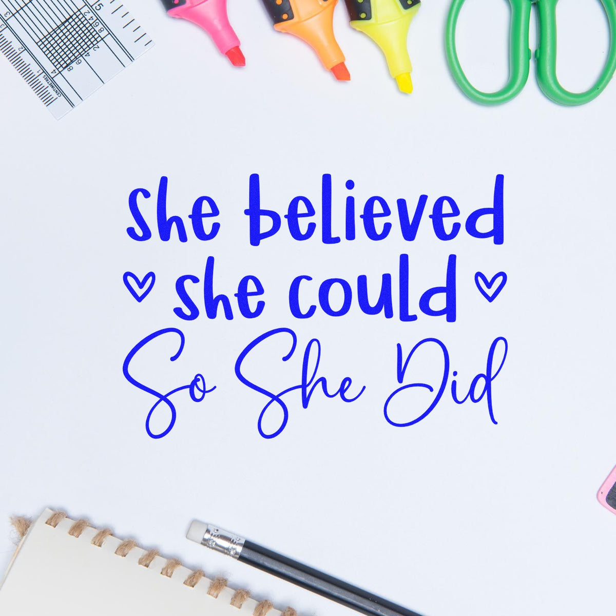 Blue stamp-and-seal quote, She believed she could so she did, with pink, orange, yellow markers, ruler, scissors, and pencil.