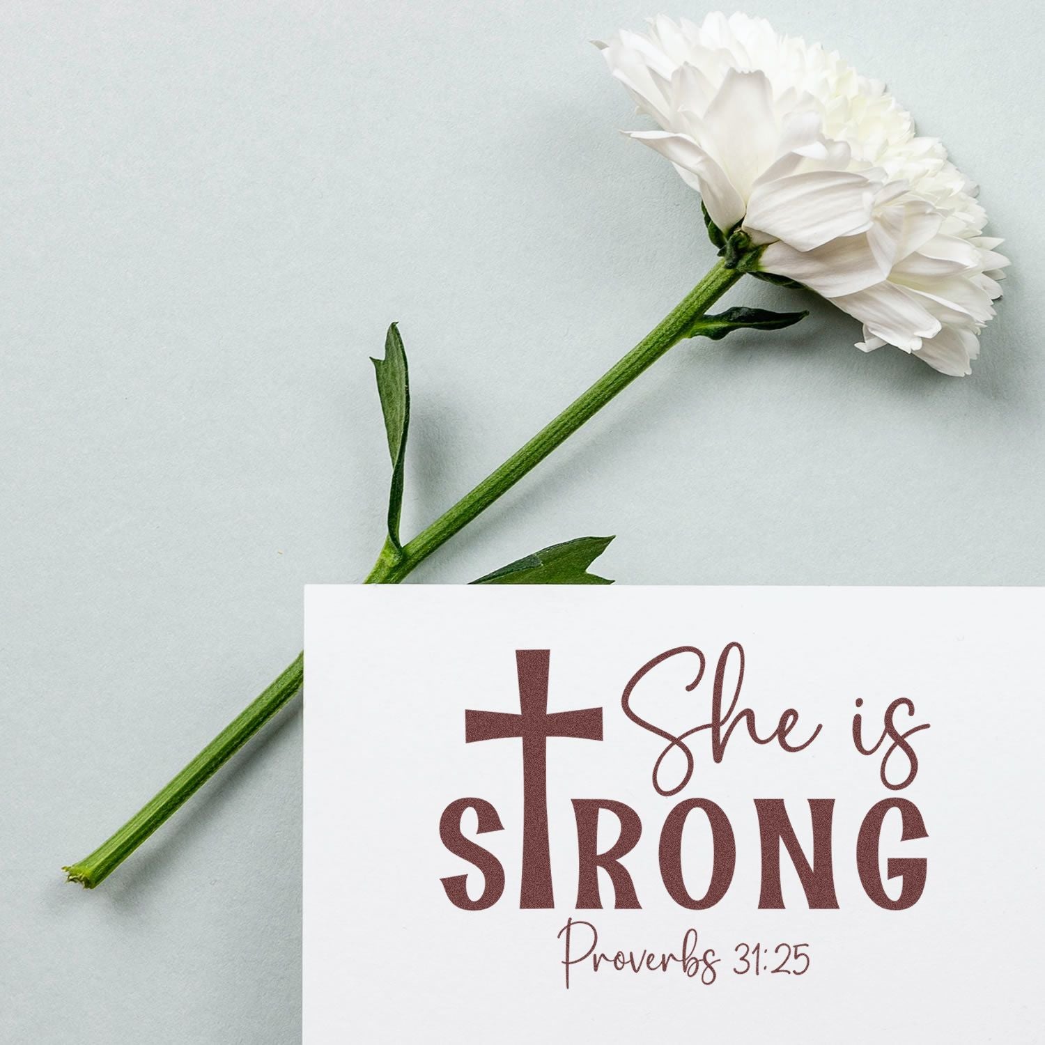 White flower beside a burgundy Christian stamp/seal reading She is Strong with cross and Proverbs 31:25.