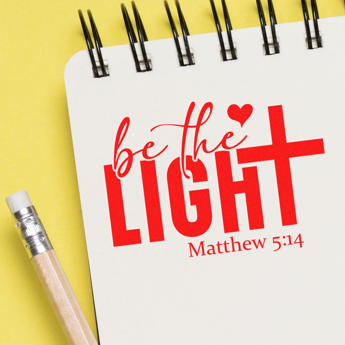 Red Christian stamp and seal design reading be the LIGHT Matthew 5:14 on a white spiral notebook, with pencil on yellow background.