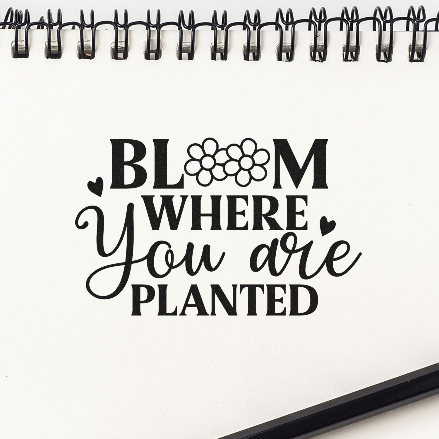 Black Bloom Where You Are Planted stamp and seal design with hearts, flower accents, on a white spiral notebook.