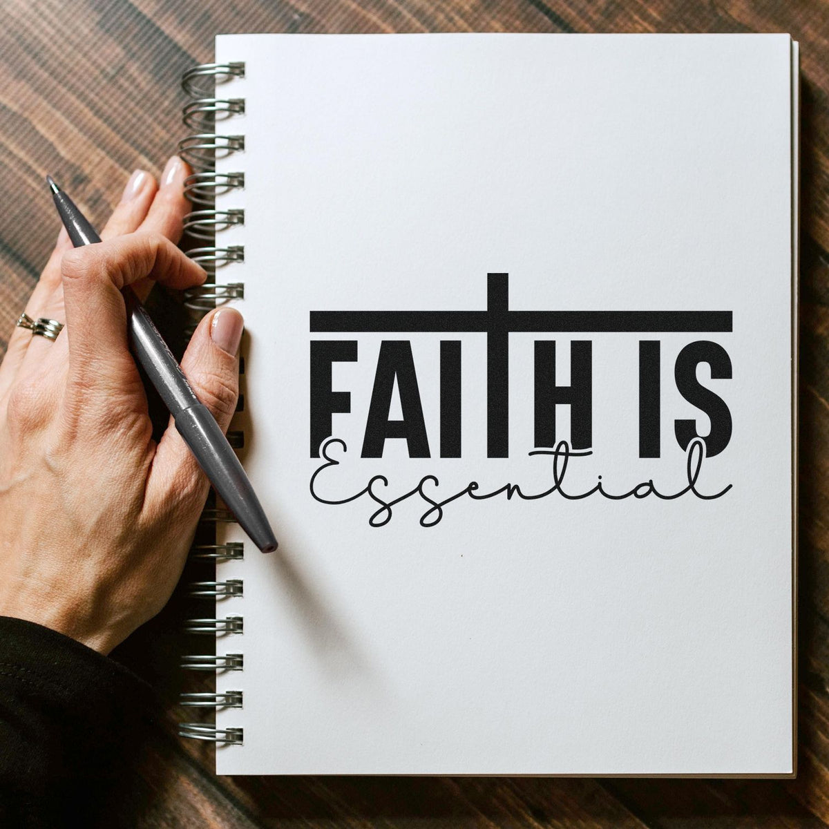 Black Faith is Essential stamp and seal on a white spiral notebook, held by a hand with a pen on a brown plaid background.