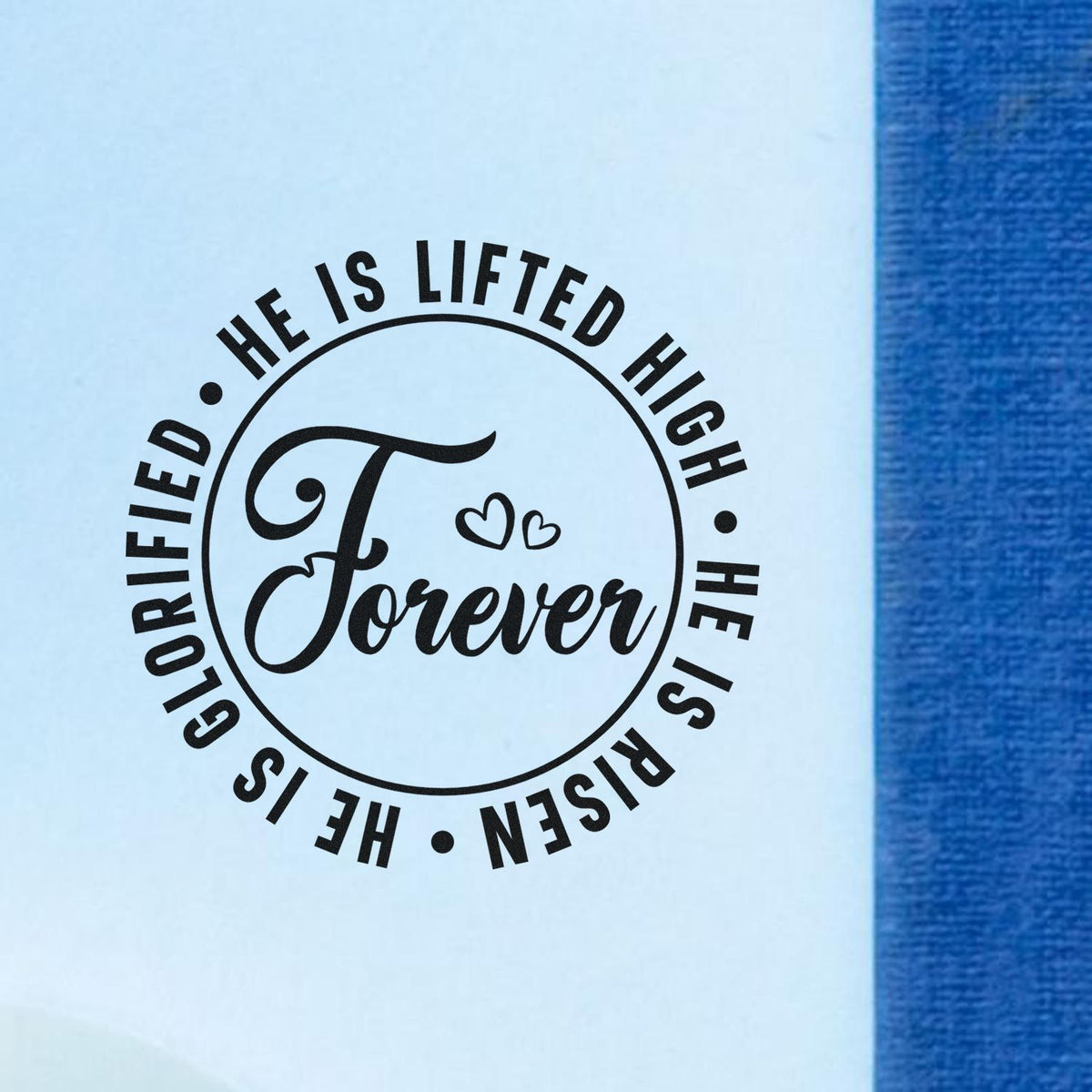 Black circular stamp seal reading He is lifted high and Forever with hearts on light blue paper beside blue fabric.