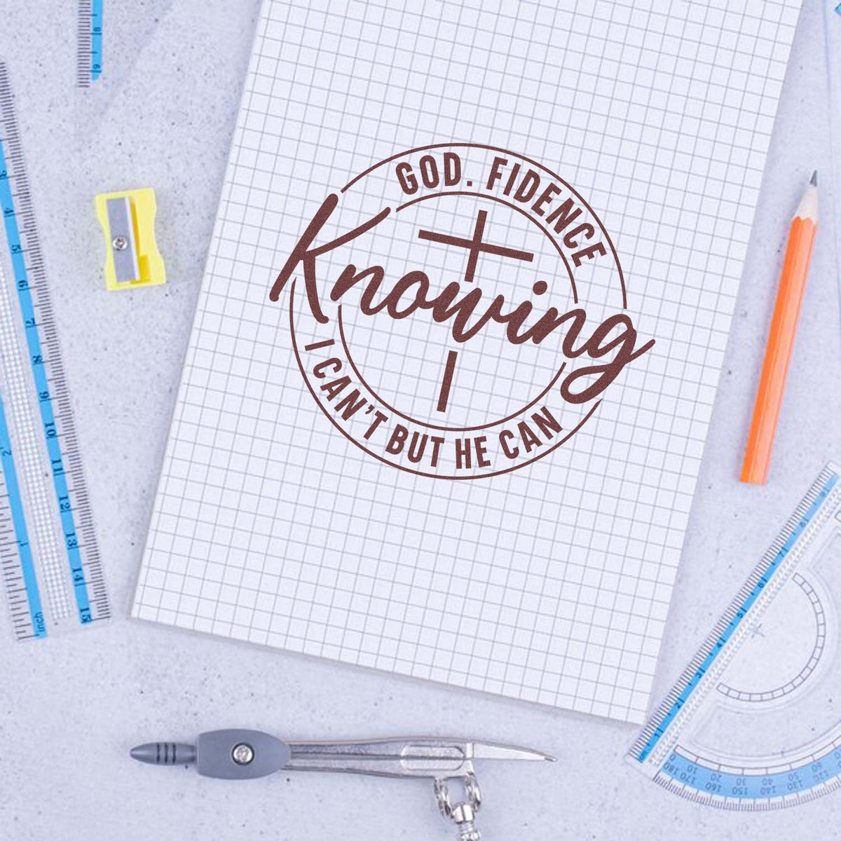 Brown faith stamp seal with cross on white graph paper, surrounded by blue rulers, orange pencil, compass, and sharpener.