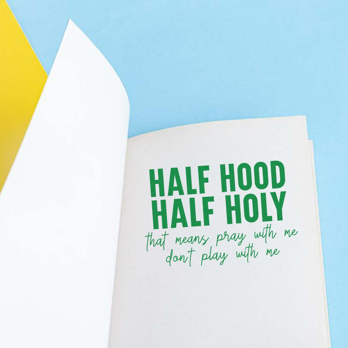 White stamp and seal product with green Half Hood Half Holy lettering on light blue background and yellow accent.
