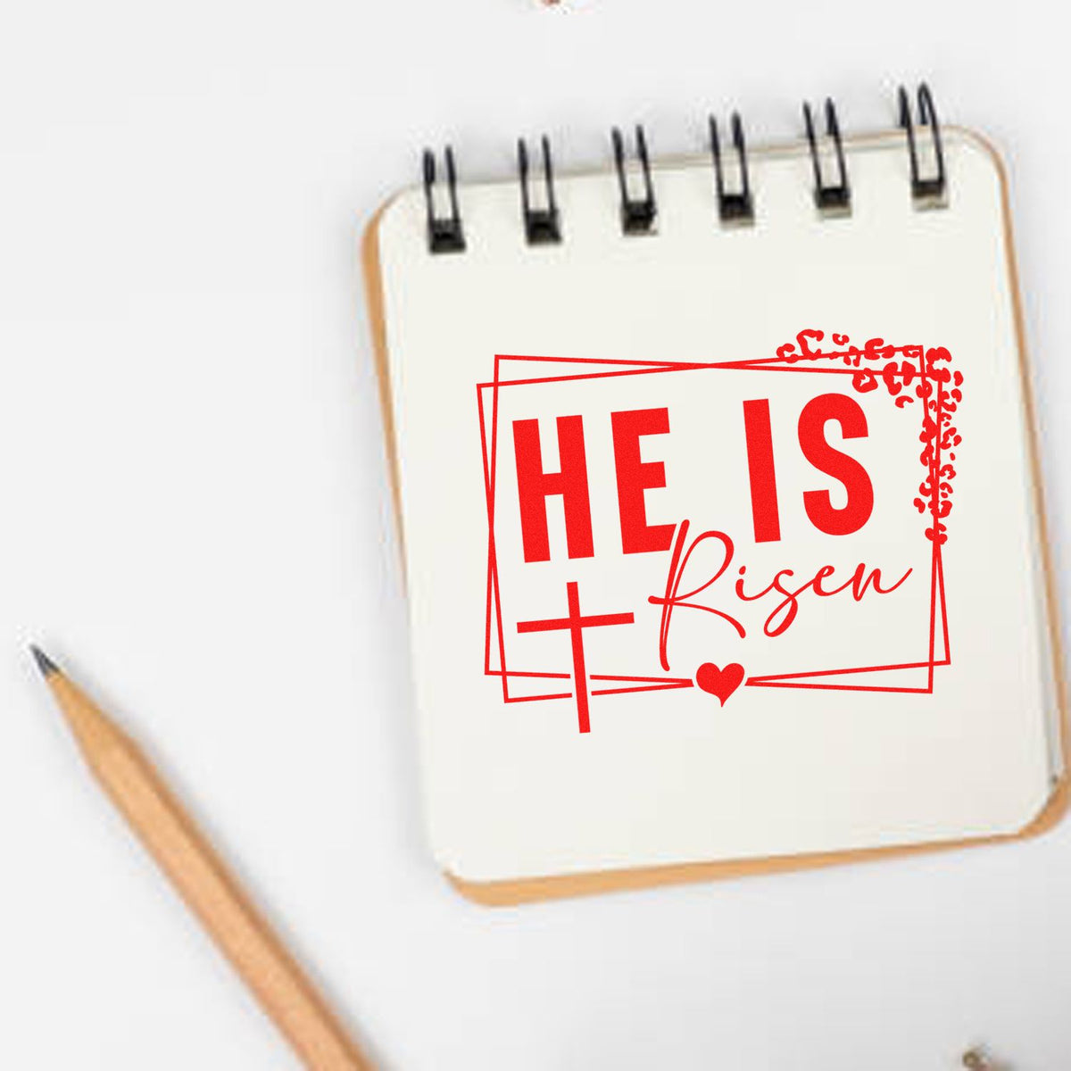 Red Christian stamp and seal reading HE IS Risen on a white spiral notebook, with a pencil beside it.