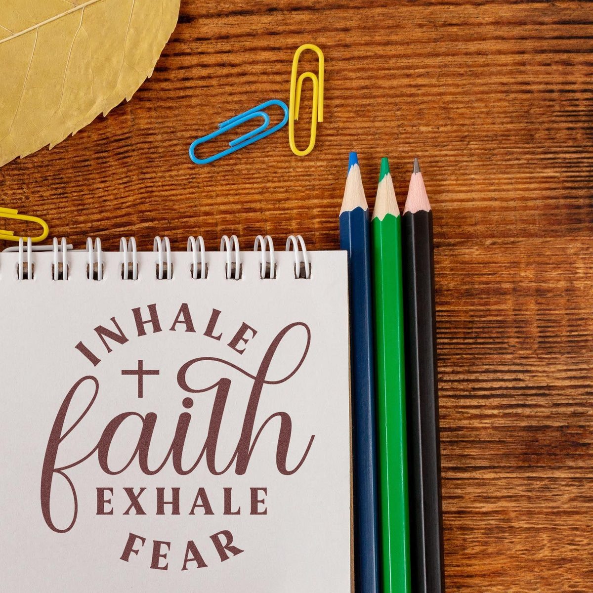 Faith-themed custom stamp and seal on a spiral notebook, beside blue, yellow, green, and black pencils on a wood desk.