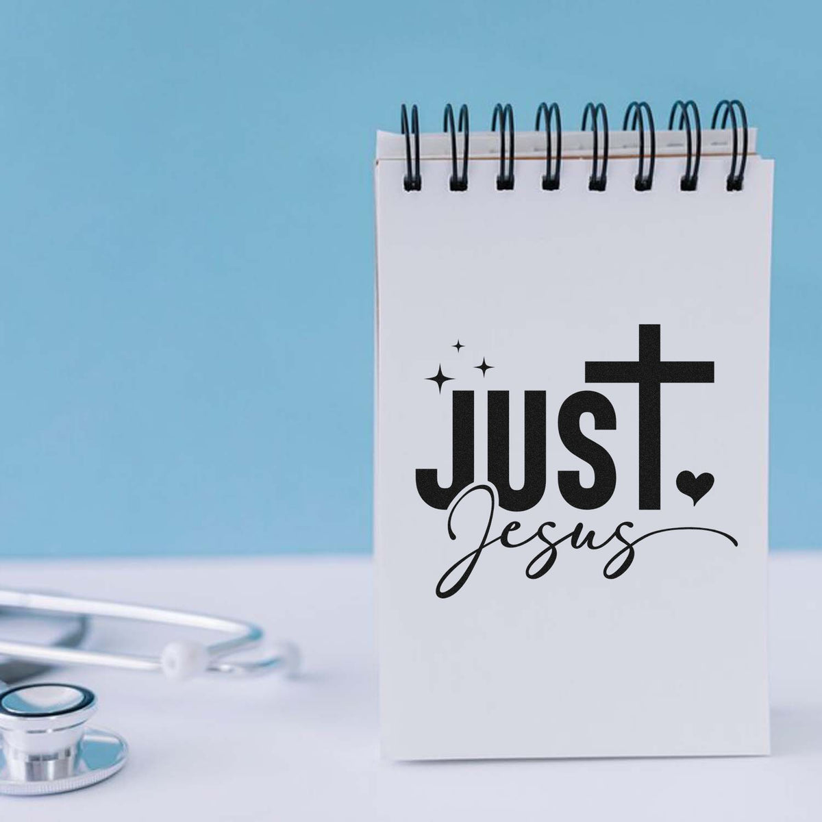 White spiral notebook with black Just Jesus stamp and seal design beside a silver stethoscope on a light blue background.