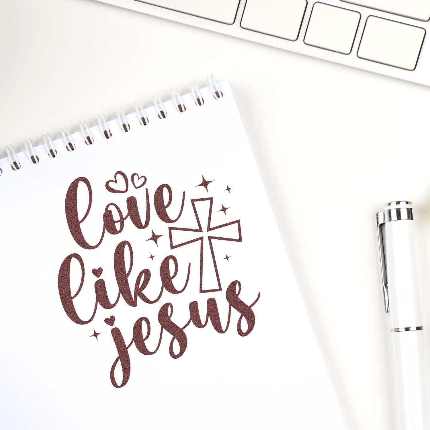 Brown Love Like Jesus faith stamp/seal design with hearts and cross on a white spiral notebook, beside a white pen.