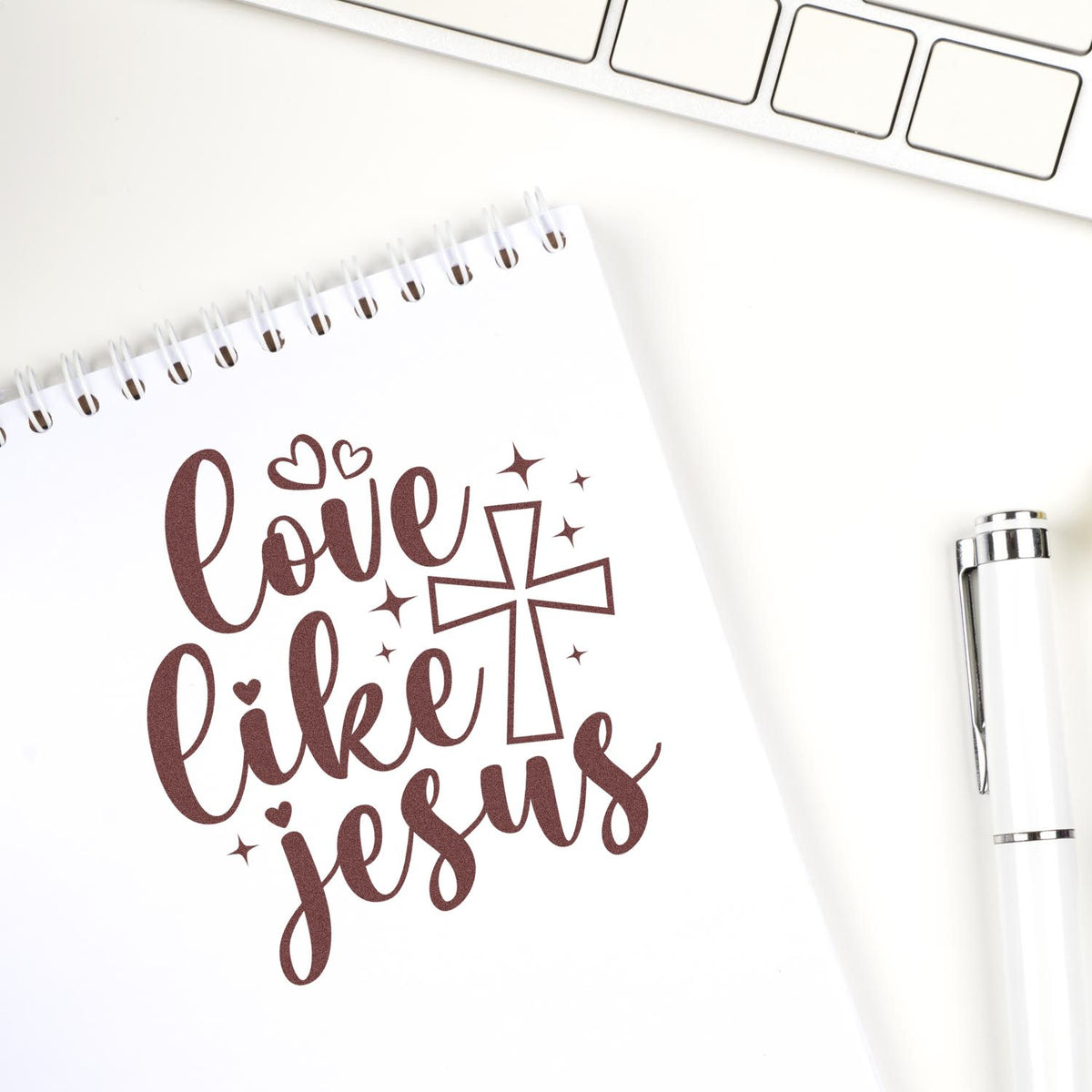Brown stamp imprint reading love like jesus with hearts and a cross on a white spiral notebook, beside a white pen.
