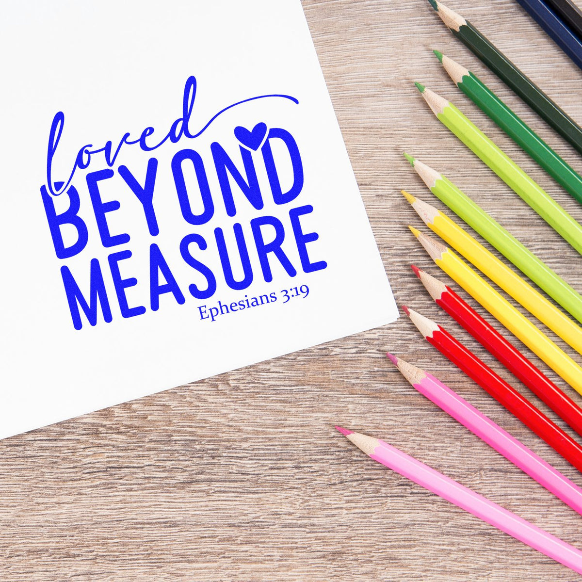 Blue scripture stamp/seal reading Loved Beyond Measure with Ephesians 3:19, beside colorful pencils on a wood desk.