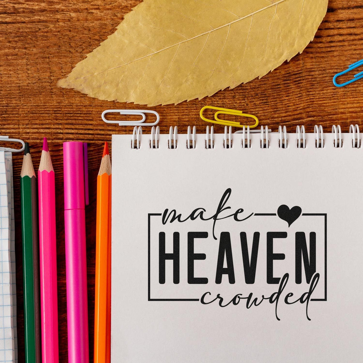 White spiral notebook featuring a black custom stamp reading make HEAVEN crowded, with colorful pencils and paper clips.