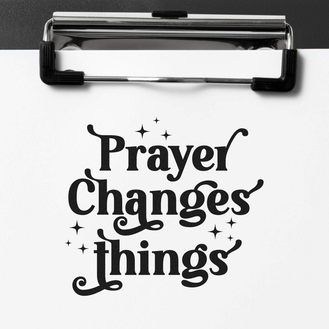 Black decorative stamp/seal reading "Prayer Changes Things" with sparkles on white paper, clipped to a silver clipboard.