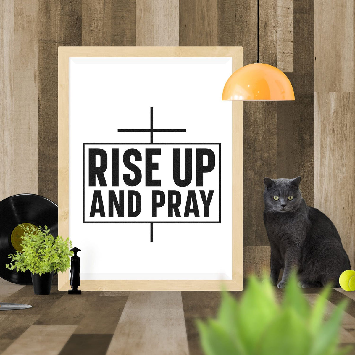 Rise Up and Pray stamp and seal design in black on white, framed in light wood against a warm wood wall with orange lamp and gray cat.
