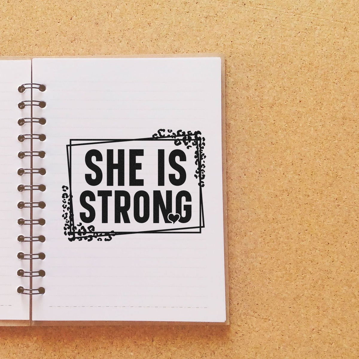 Black decorative stamp seal reading SHE IS STRONG on a white notebook page against a tan cork background.