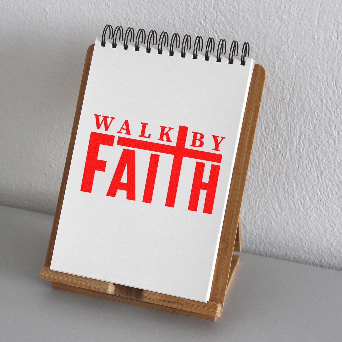 White spiral notepad on wooden stand featuring bold red WALK BY FAITH stamp and seal design
