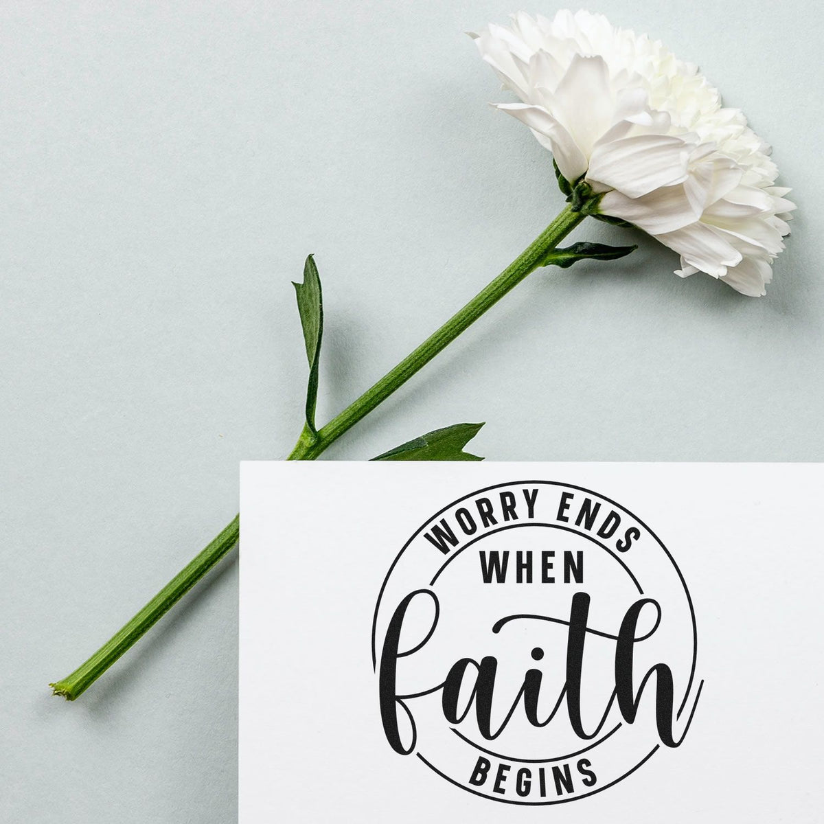 White flower beside black faith stamp seal on pale gray background, reading Worry ends when faith begins.