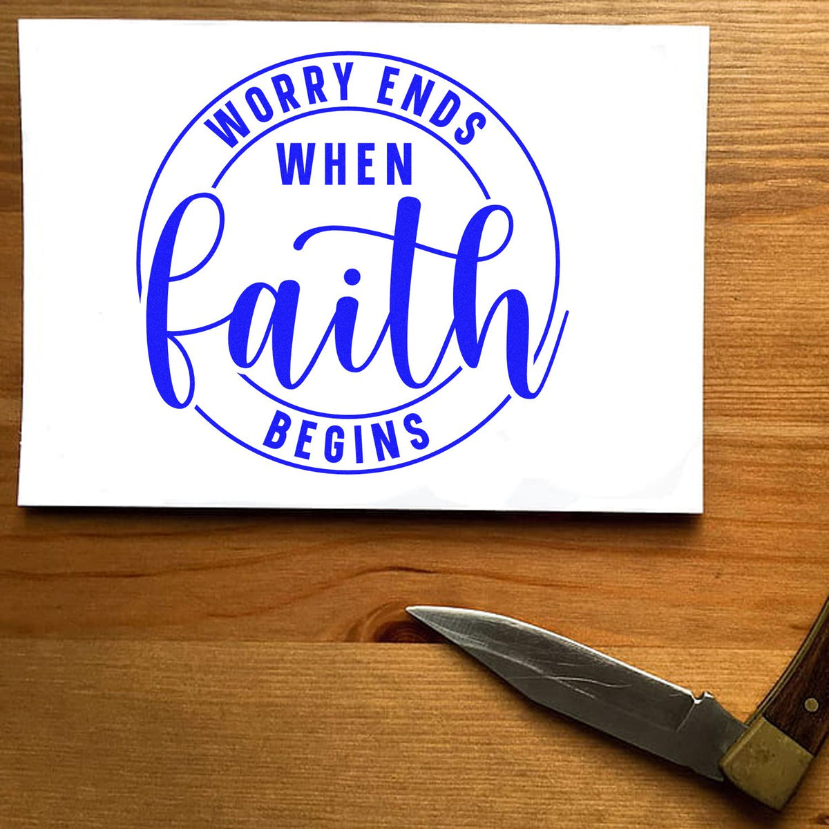 Blue faith stamp seal on white card, circular slogan Worry Ends When Faith Begins on a wooden table.