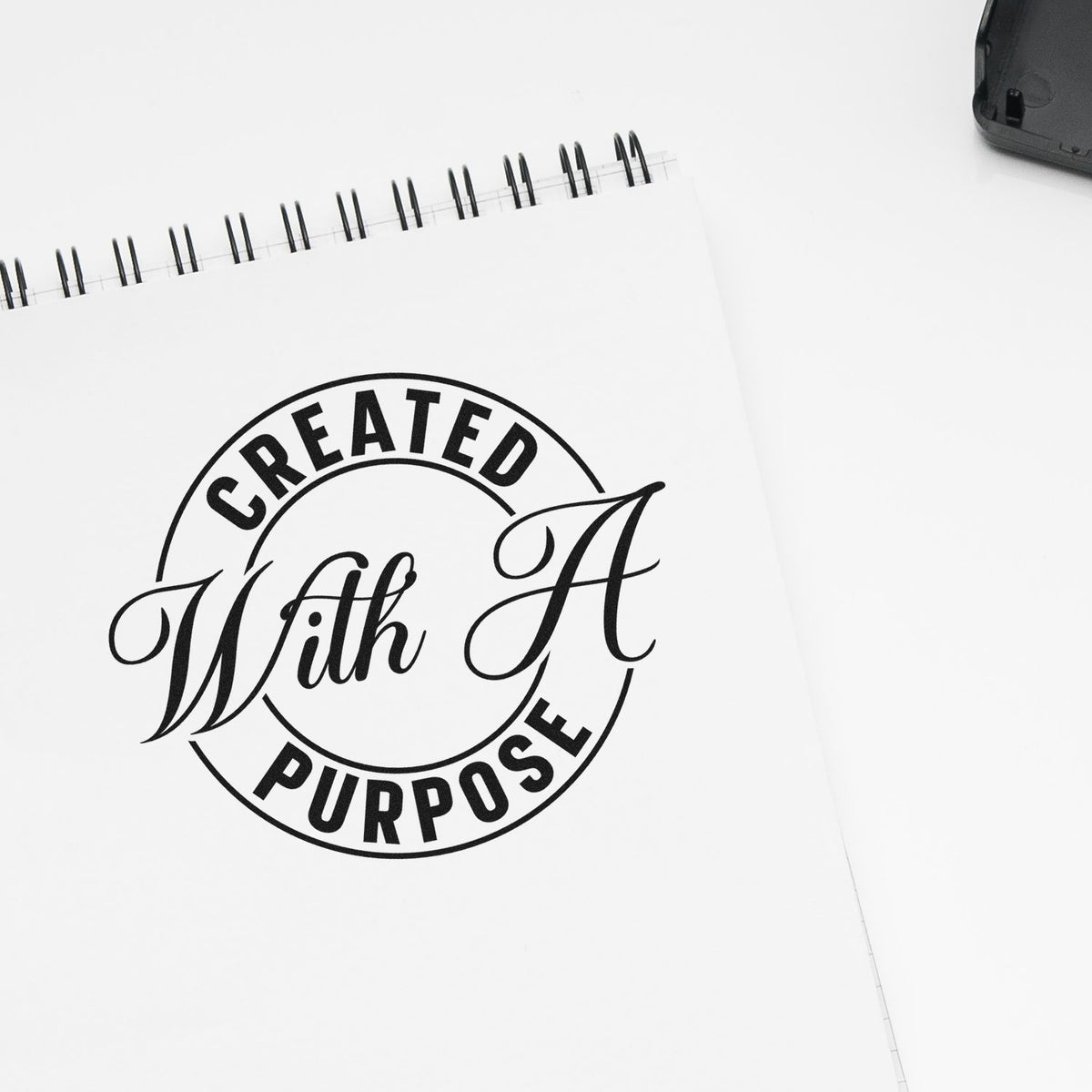 Black Created With A Purpose stamp and seal design on a white spiral-bound notebook, featuring a circular logo.