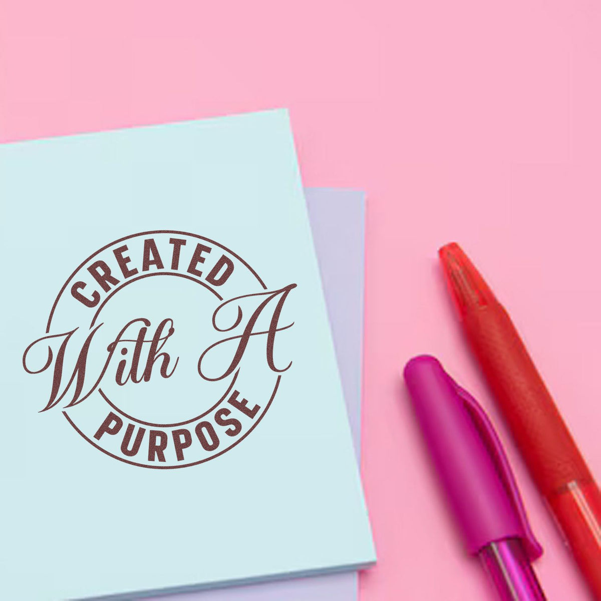 Custom brown circular stamp seal reading Created With A Purpose on light blue card, beside red and magenta pens on pink background.