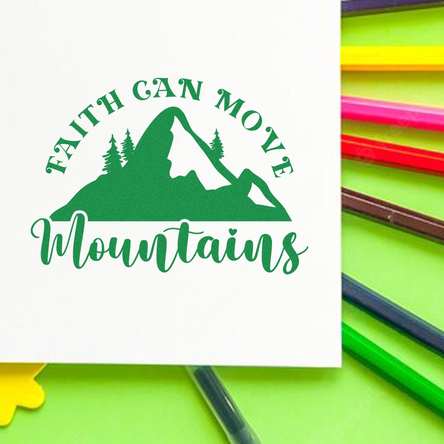 Green custom stamp seal with mountain logo and Faith Can Move Mountains on white, beside colorful pens.