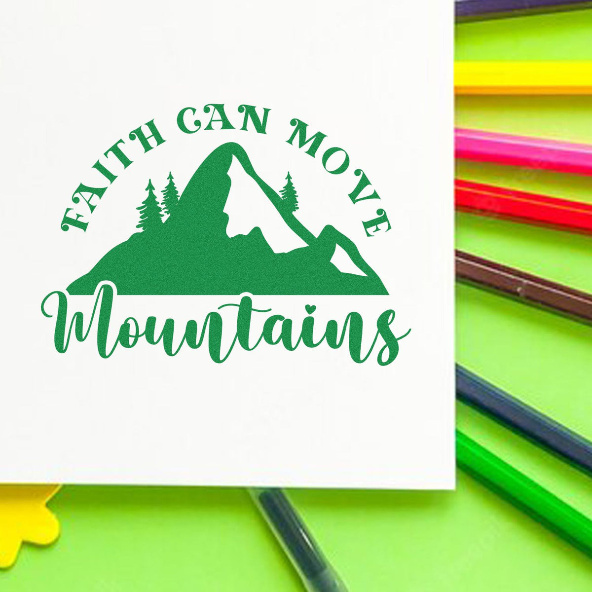 Green faith-can-move-mountains stamp seal with mountain and pine tree design on white, beside colorful pens.