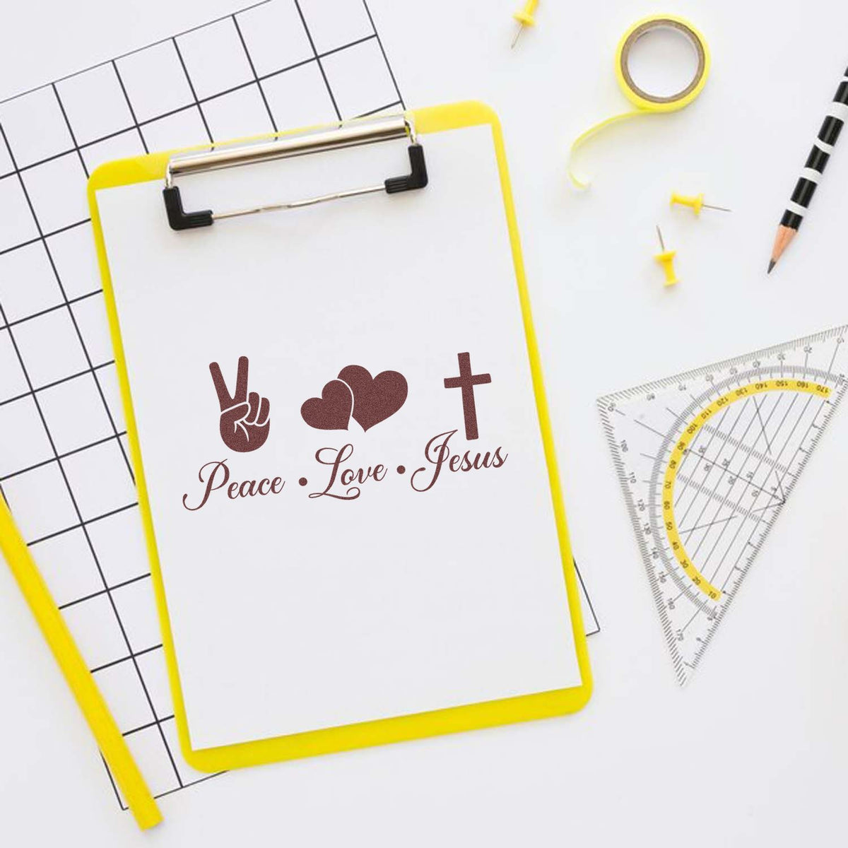 Yellow clipboard stamp with brown Peace Love Jesus design, peace sign, hearts and cross, on white desk with rulers and pencil.