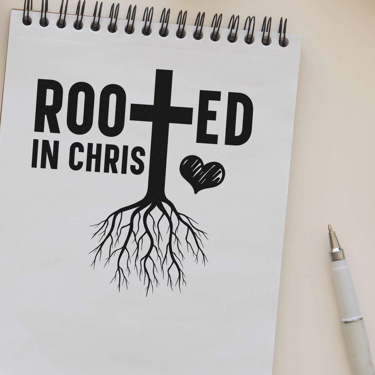 Black stamp and seal design reading Rooted in Christ with a cross, roots, and heart on a white spiral notebook.