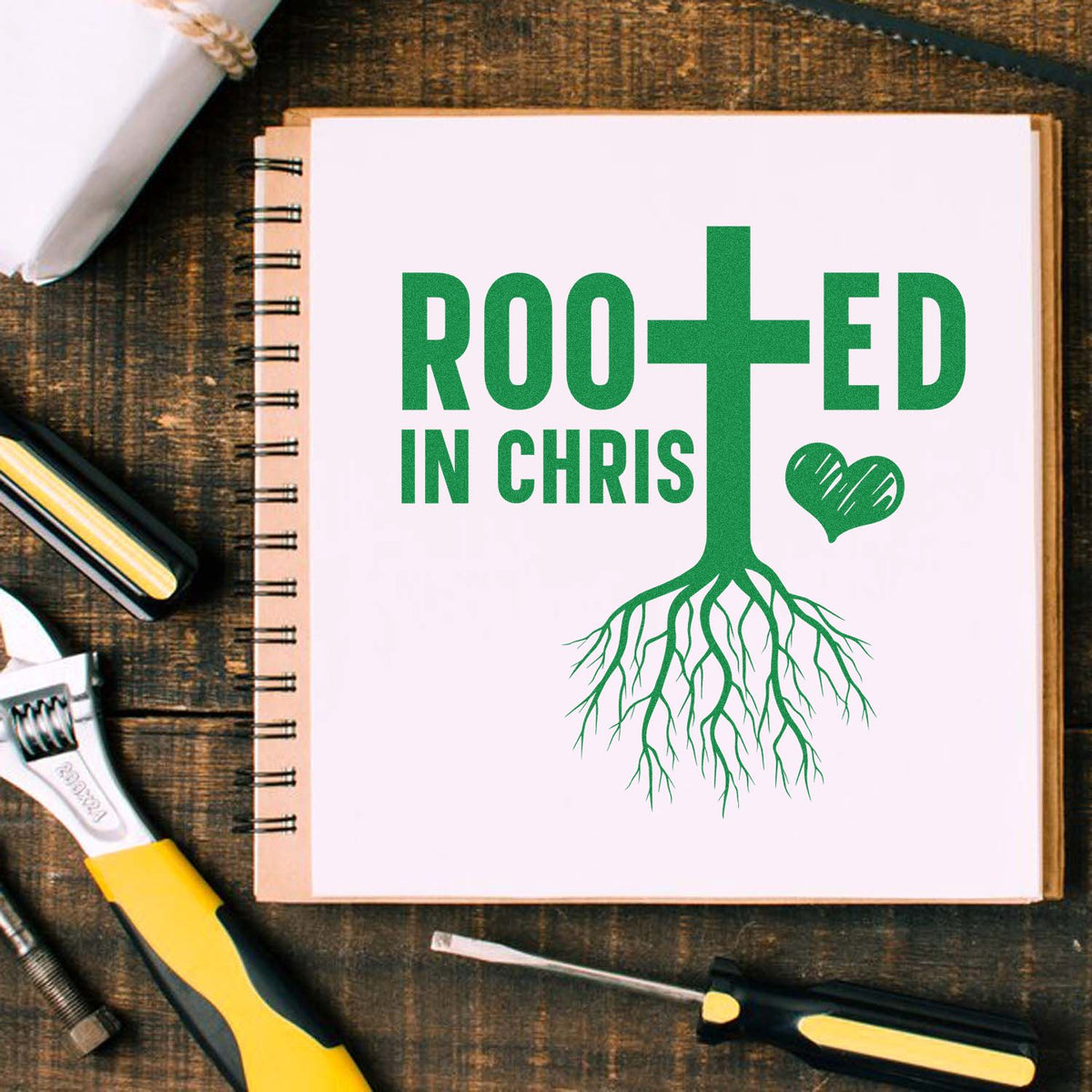Green Rooted in Christ stamp and seal design with cross, heart, and roots on a pink notebook, surrounded by tools.