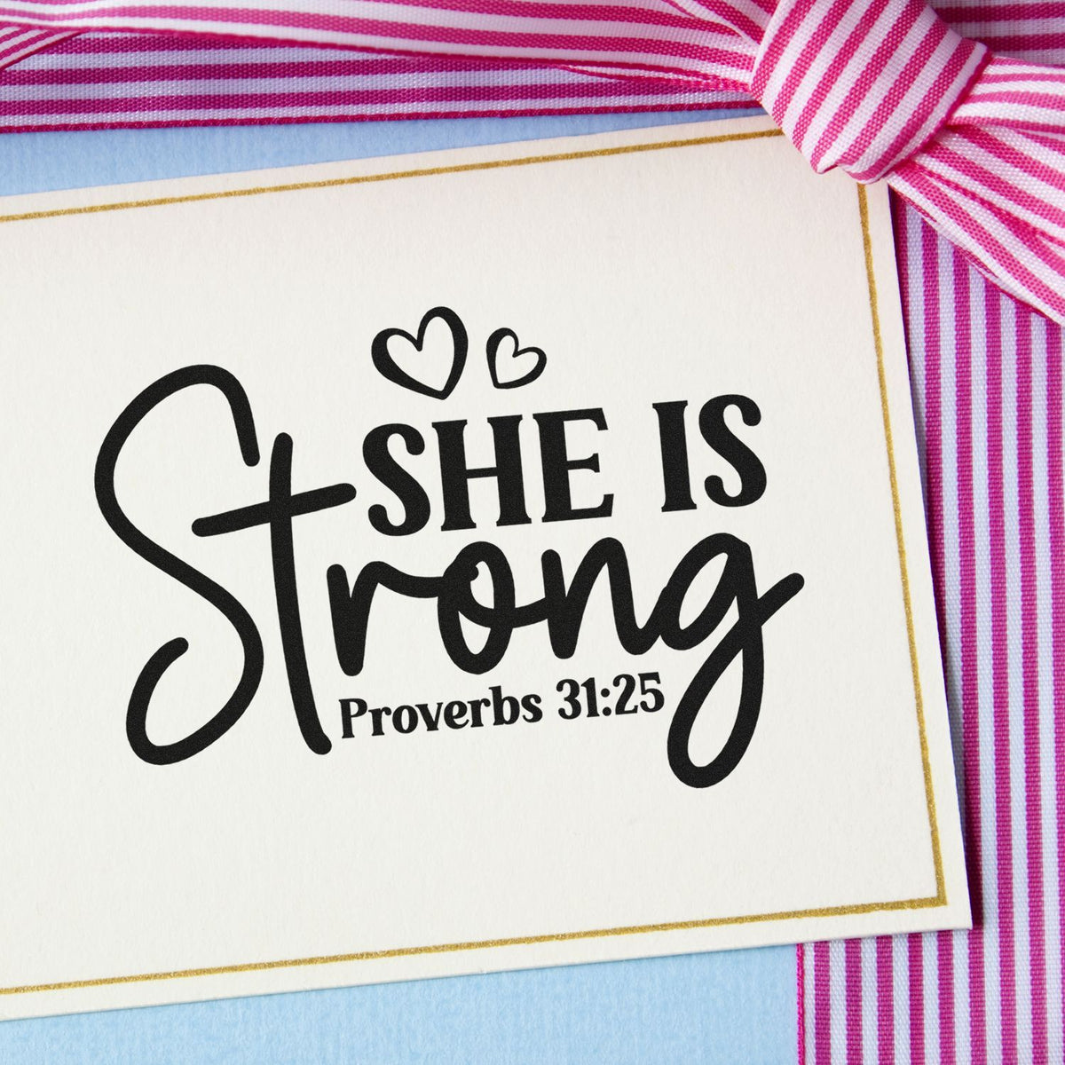 Black She Is Strong stamp and seal with hearts and Proverbs 31:25 on white cardstock, pink striped ribbon.