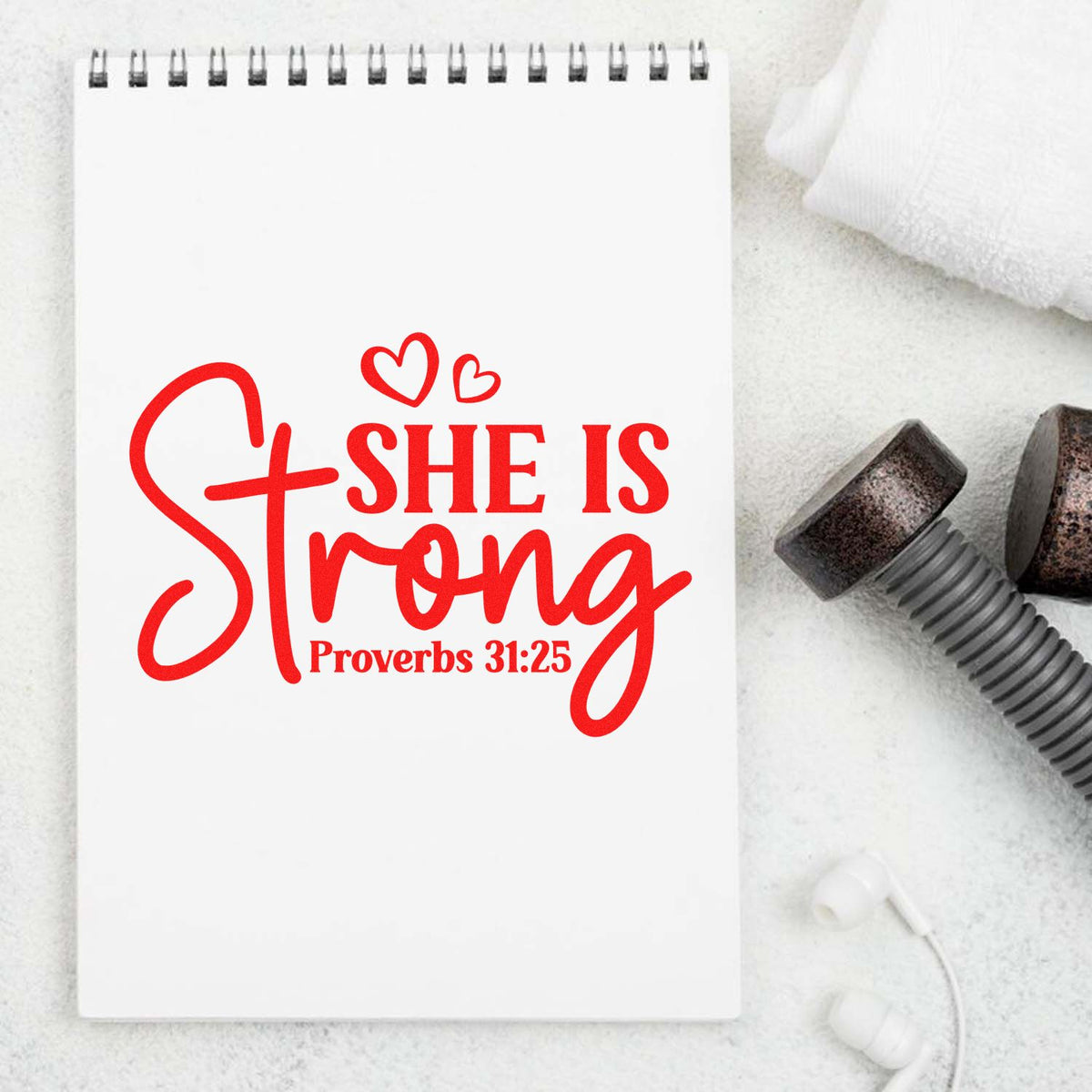White spiral notebook with red She is Strong Proverbs 31:25 text, hearts, and black stamp and seal tools on a soft white background.