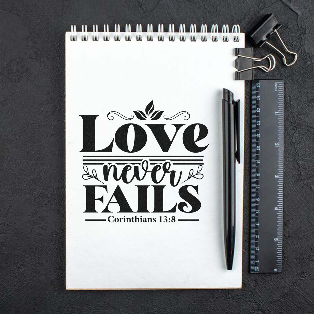 Black custom stamp seal reading Love never fails with Corinthians 13:8 on white notebook, styled with ruler and pen.